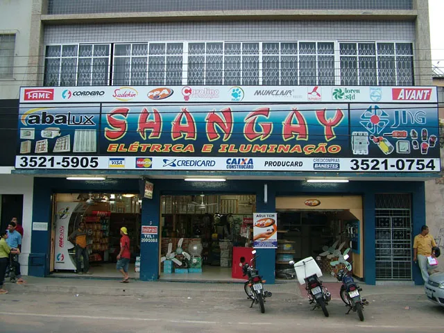 Fachada da loja: no letreiro azul, Shangay em letras vermelhas, Elétrica e Iluminação em amarelo, os telefones 3521-5905 e 3521-0754 e logotipos de marcas; embaixo, três portas abertas e motos paradas na calçada