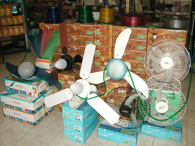 Ventiladores de teto, com pás de várias cores, e ventiladores com grade expostos sobre as caixas; no alto da pilha, cinco carretéis coloridos